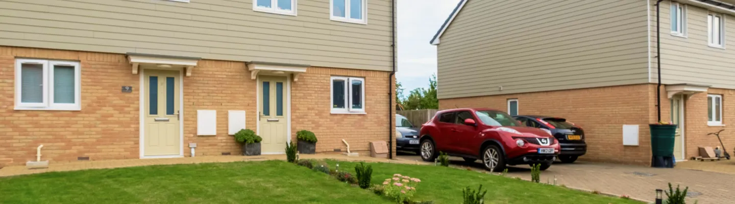 Row of two-storey beige-and-brick houses with small front gardens and driveways; parked cars, a scooter and bins visible.