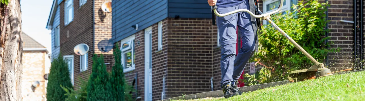 Person in blue clothing using a grass trimmer to edge a sloped lawn beside brick houses; a tree trunk and shrubs frame the scene.
