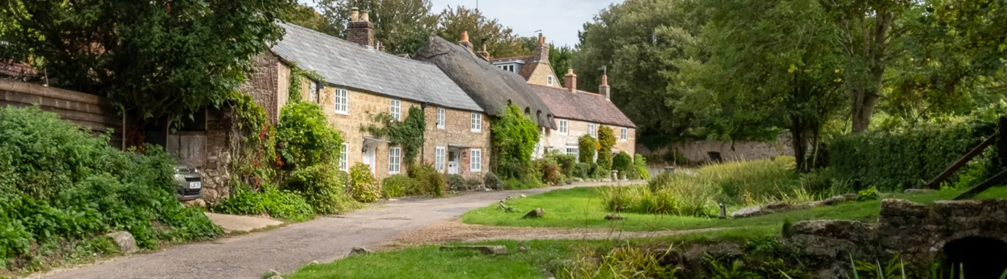 Row of stone cottages with slate roofs along a village street, ivy-covered walls and a small stone bridge over a grassy bank on the right.