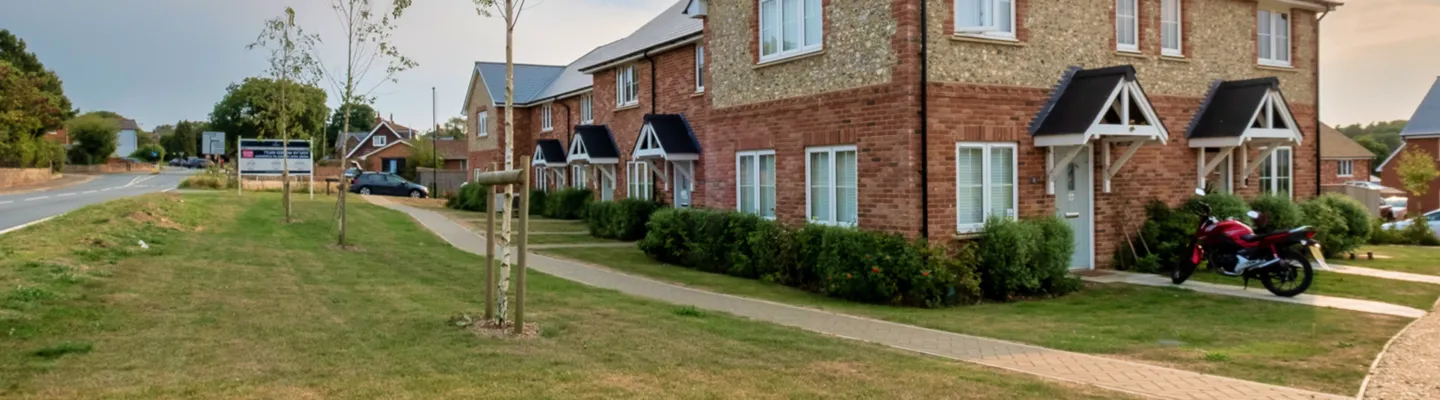 Suburban street with red-brick houses and hedges; a red motorcycle sits by a front door. A curved path runs along lawns, with road in the distance.