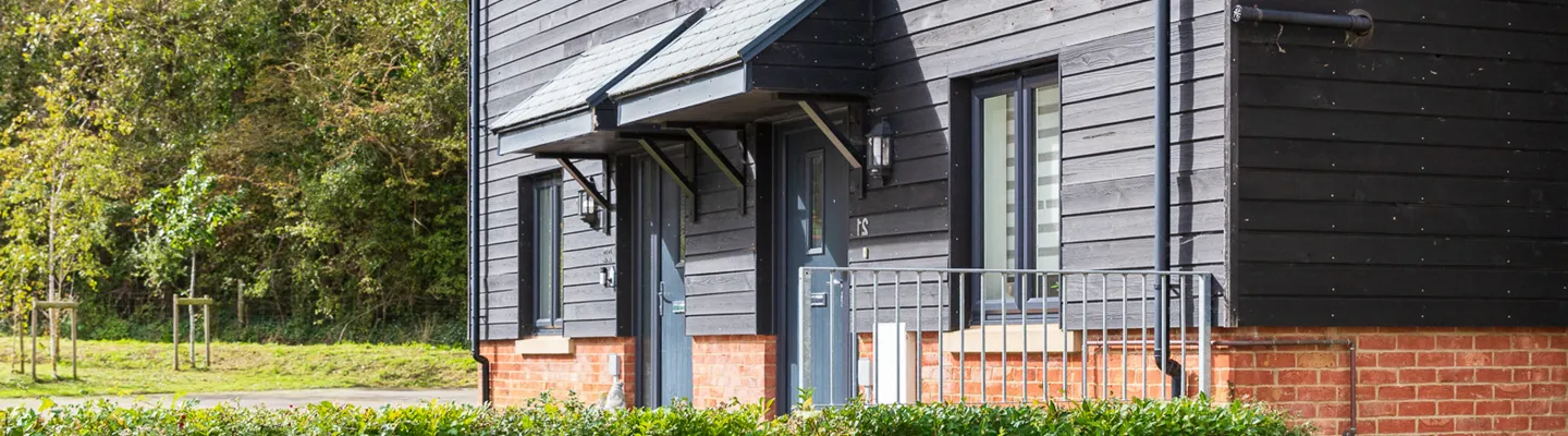 Row of modern houses with dark wood siding and brick bases, doors under canopies, railings on brick fronts, with trees and grass to the left.