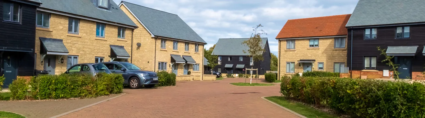 Row of two-storey houses with stone and dark timber facades, a curved brick road, hedges, and parked cars along a sunny residential street.