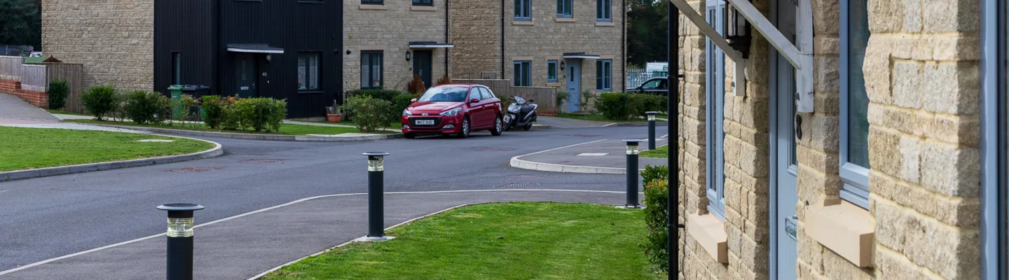 Wide residential street with light stone houses, a red car parked by the curb, a scooter behind it, and black bollards along a grassy verge.