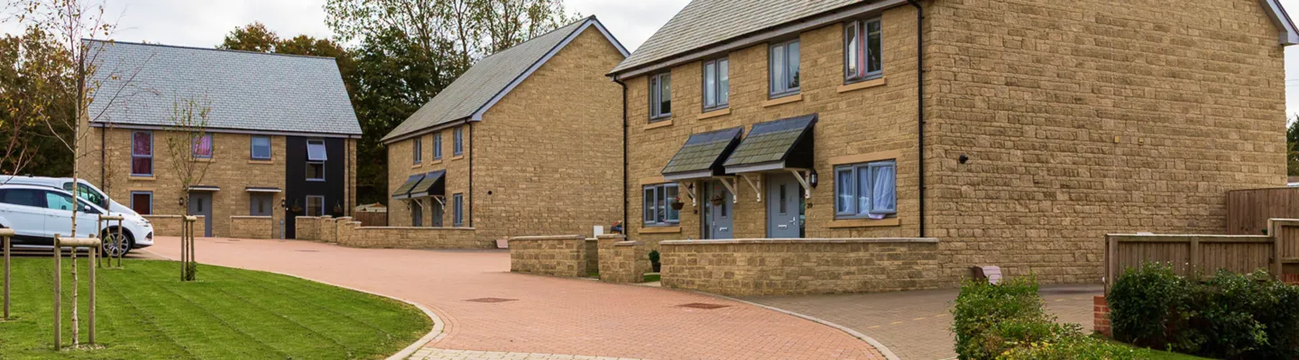 Row of beige stone houses with slate roofs along a curved brick road; a white van parked left, young trees, and a neat lawn with wooden fencing.