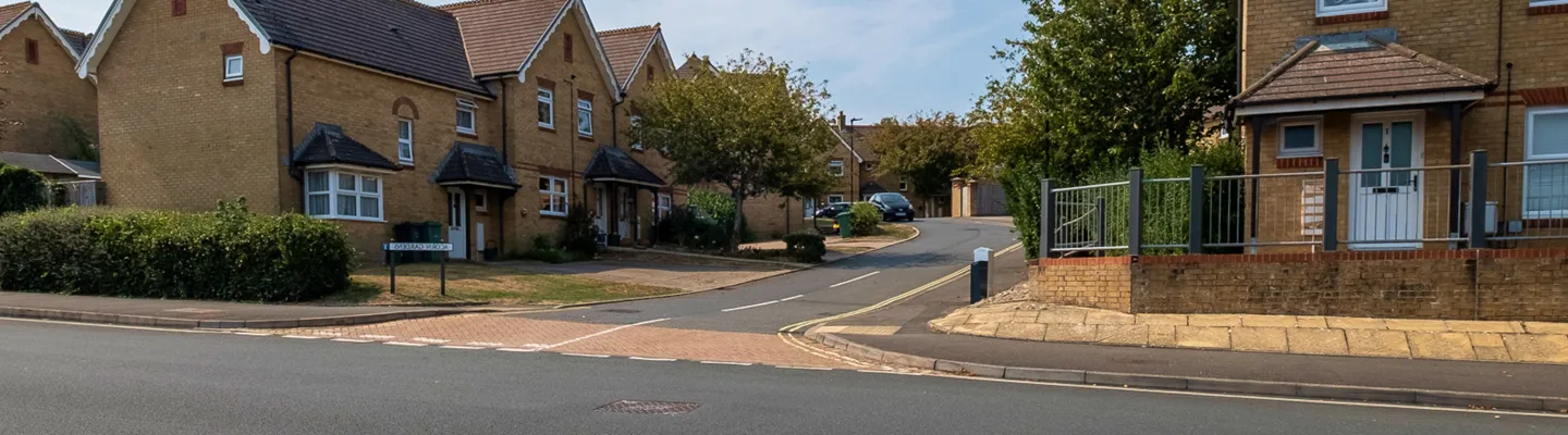 Residential street with brick houses, hedges and a parked car on the left; a curved road leads past a raised-front home with a metal railing on the right.