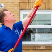 A person in a blue polo shirt and orange gloves uses a long red pole against a brick wall, reaching up toward a white-framed window.