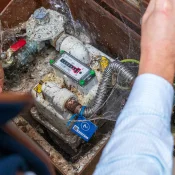 A person in a blue striped shirt works in a brick-lined outdoor utility pit, handling a valve and meter among pipes, with a blue tag and cobwebs.