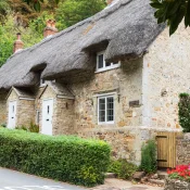 Thatched stone cottage with white doors, hedged front, and a stone wall; a car on the road's left and red flowers near a wooden gate.