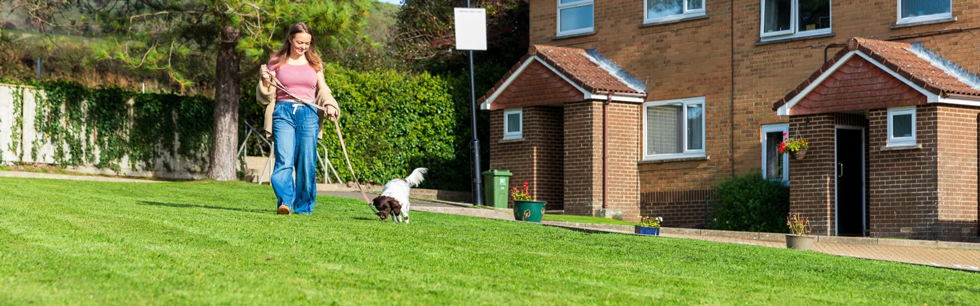 A person in a pink top and blue jeans walks a small brown-and-white dog on a leash across a green lawn by a brick house with a porch and potted plants.
