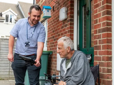 A person in a striped shirt with a lanyard shows a tablet to an older adult in a wheelchair outside a brick house, with a blue birdhouse on the wall.