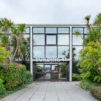 Front view of Building 41 with a glass facade and central entry, flanked by palm trees and lush shrubs along a paved walkway.