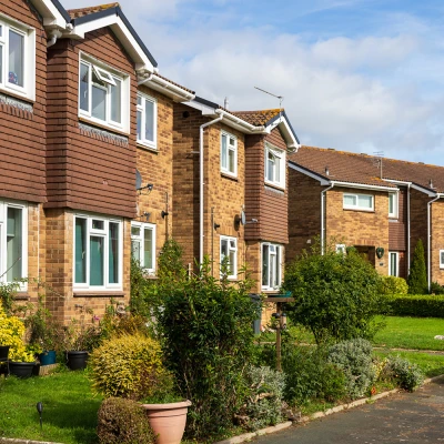 Row of red-brick terraced houses with white-framed windows and small front gardens filled with plants; a lamp post and a clear blue sky.