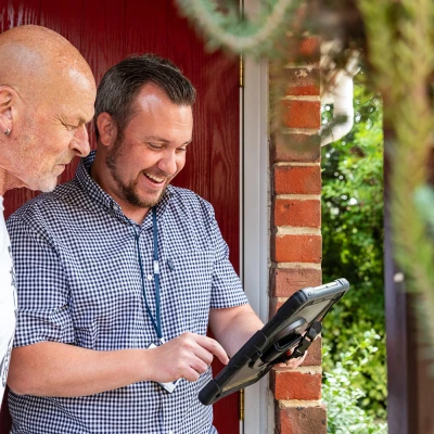 Two adults stand by a red door, smiling while looking at a rugged tablet. One wears a white T-shirt, the other a checked shirt with a lanyard; brick and greenery behind.