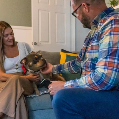 Two people sit on a grey sofa in a cosy living room, smiling at a brown bulldog wearing a red bow tie as one strokes its head.