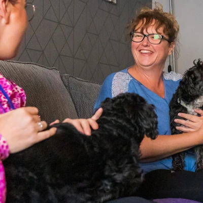 Two adults sit on a grey sofa with two dogs: a large black dog and a small black-and-white terrier held by a person in a blue top and glasses.